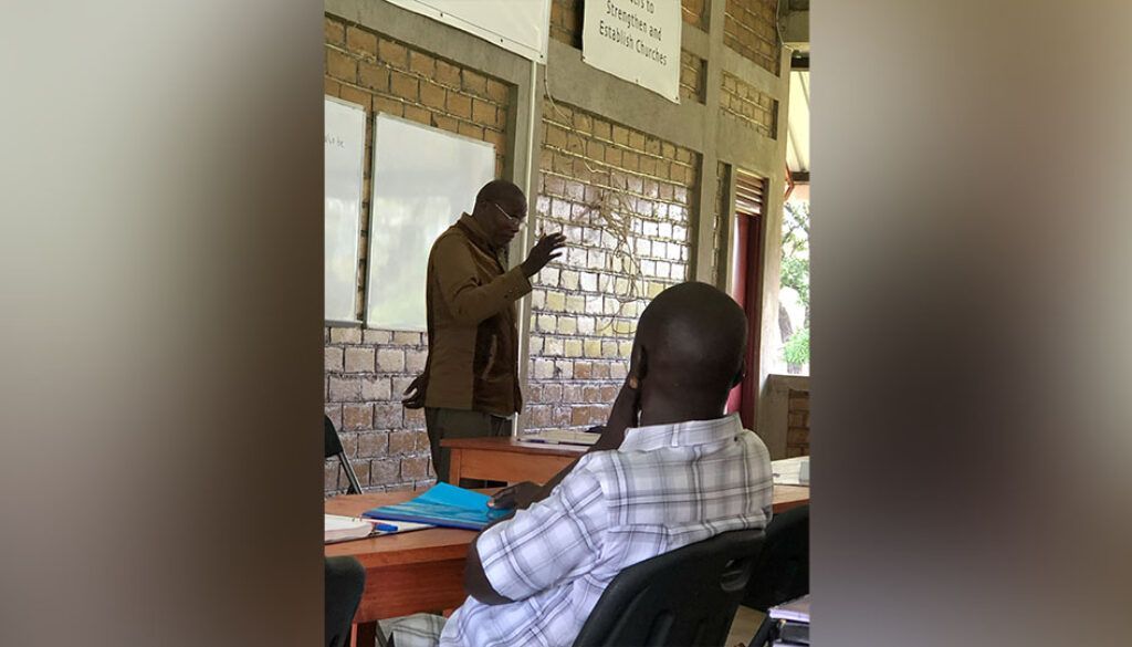 Blog 10 pic copy Man lecturing in front of a seated audience in a room. Whiteboard and papers on the wall.