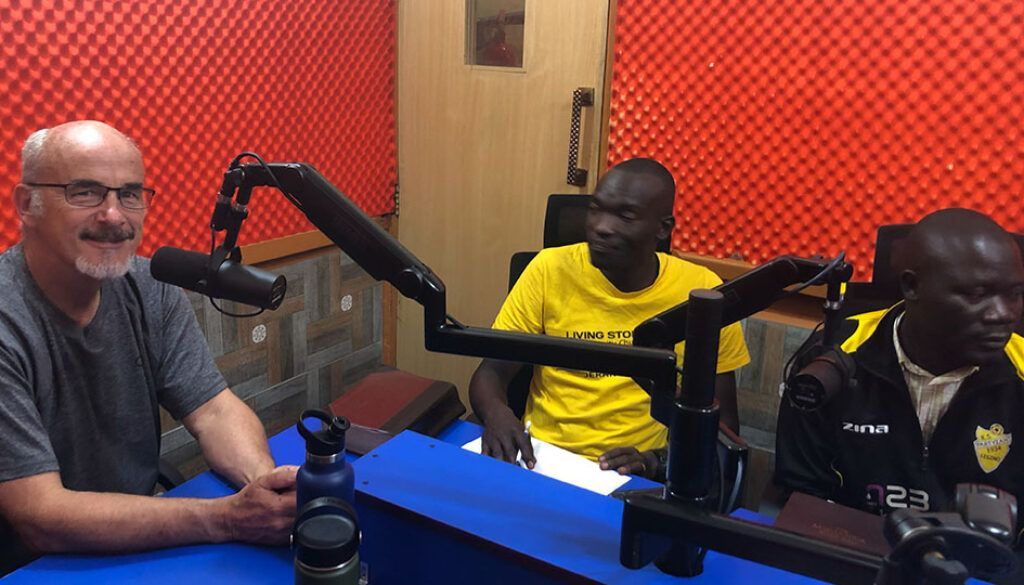 Blog 8 pic Three men at a table in a radio studio with microphones. Red and black soundproofing on the walls.