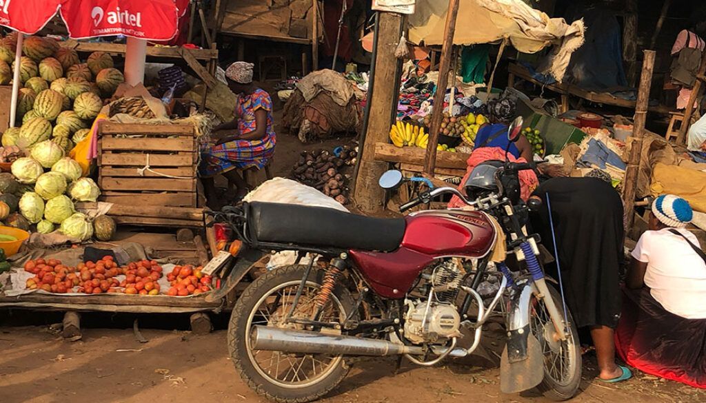 Blog 9 pic Motorcycle parked in a bustling outdoor market with food stalls, shoppers, and a red and white umbrella.