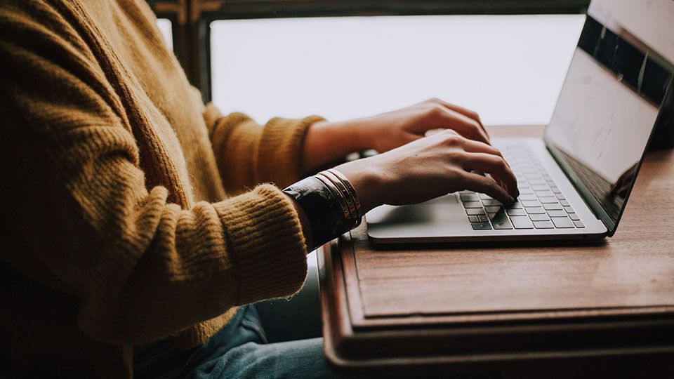 Person typing on a laptop at a wooden table, wearing a yellow sweater, near a window.