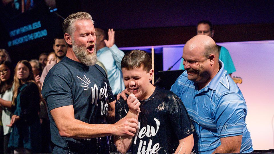 Boy being baptized in church; two men assist. People in background, dark setting.
