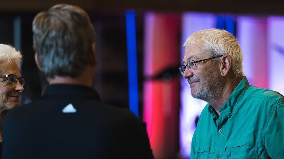 Man in green shirt smiles while talking, indoors. Others partially visible.