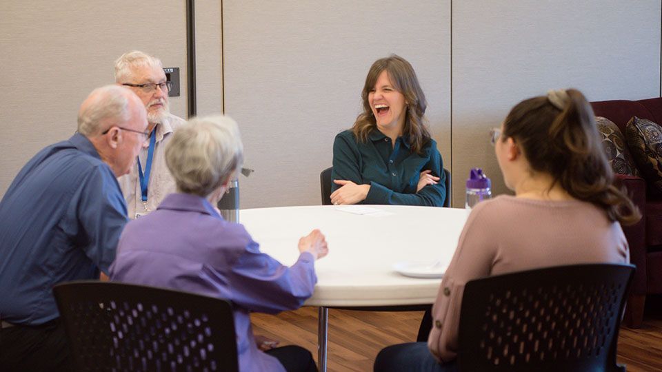 Five people seated around a table, laughing. One woman in teal shirt laughs; others look on.