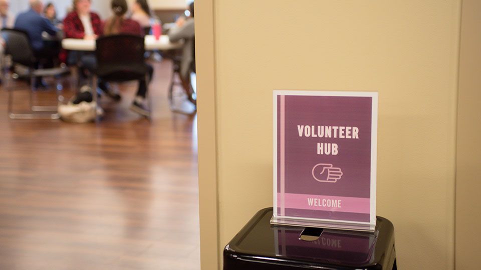 Volunteer Hub sign on a black stand in a room with a blurred background of people at tables.