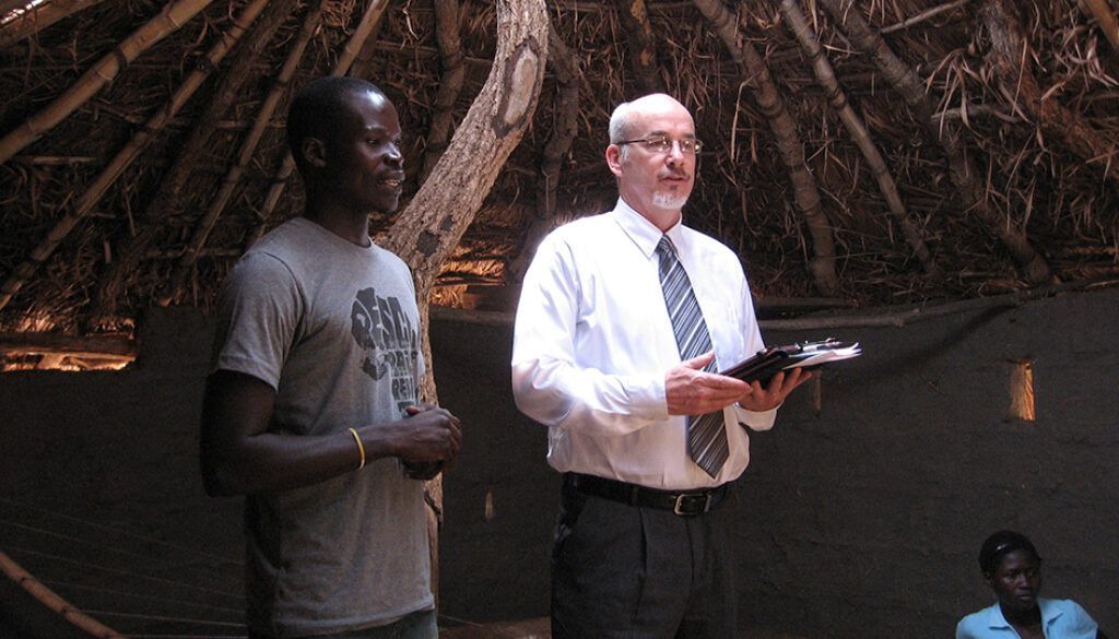 Two men stand inside a thatch-roofed structure. One in a white shirt and tie holds a book and speaks. The other man listens.