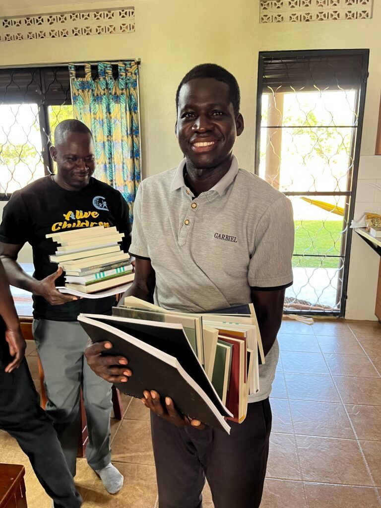 Two men holding stacks of books in a room with windows and blue curtains, smiling.