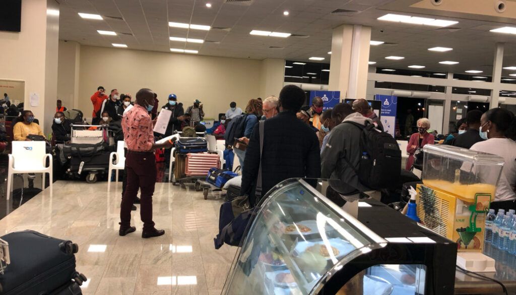 People in an airport. Some are masked, pulling luggage, others seated. A person reads from papers.