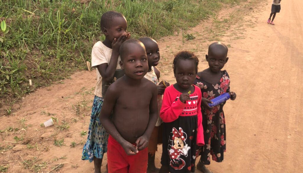 Children standing on a dirt road, some looking at the camera. Green vegetation on the left.