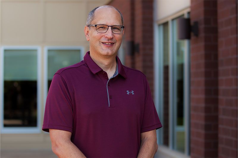 Man in maroon polo shirt and glasses smiles outside a brick building.