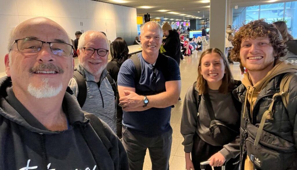 LarryImage Five people smiling at airport. Men wearing glasses and backpack, woman with a rolling suitcase, young man with curly hair.