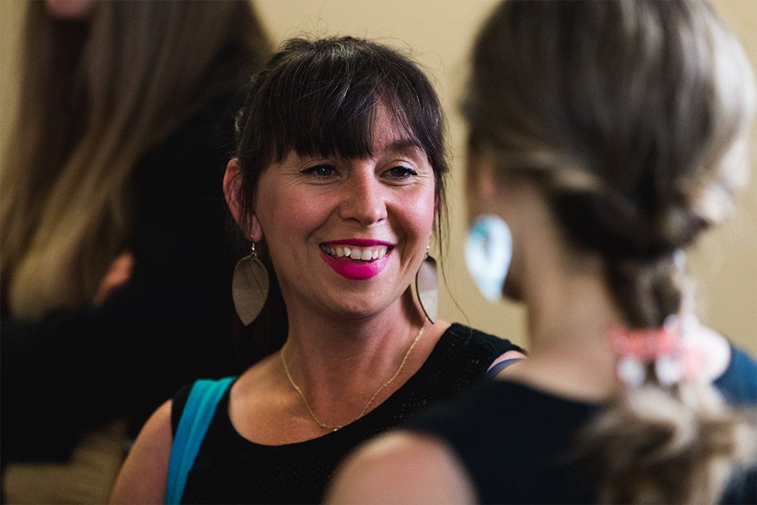 Woman with dark hair and pink lipstick smiles, talking to another woman.