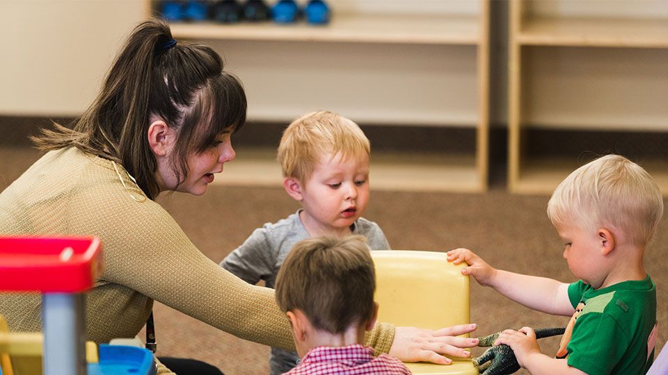 Woman helping two toddlers play with a yellow block, other children nearby in a playroom.