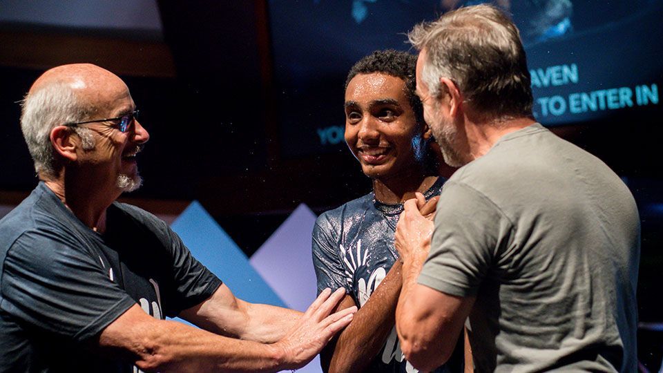 Two men baptizing a smiling person, possibly a teenager, in a church setting.