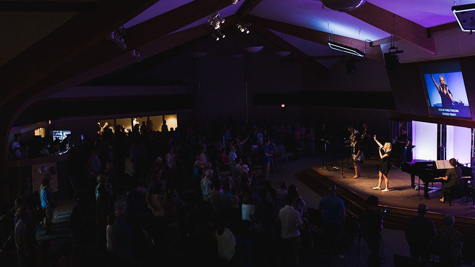 People singing in a dimly lit church with a band on stage, spotlights, and a screen.
