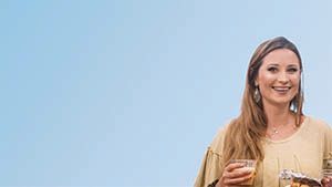 Woman smiling, holding a drink, light-colored clothing, against a blue background.