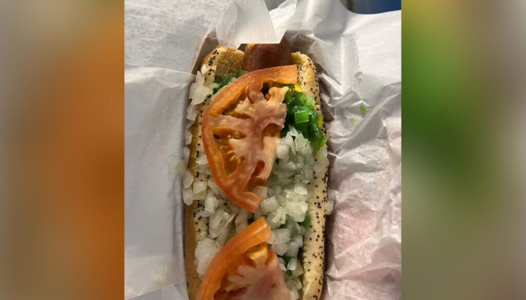 Hot dog in a bun, topped with tomato slices, diced onions, and green sauce, wrapped in white paper.