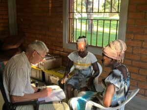 Man in light shirt, seated at a desk, talks to two seated women under a window.