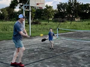 Man and child playing pickleball on a concrete court outdoors, near a basketball hoop.