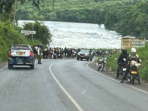 Roadside scene: Cars, motorcycles, and crowd gather near a rushing river. Green foliage and signposts visible.