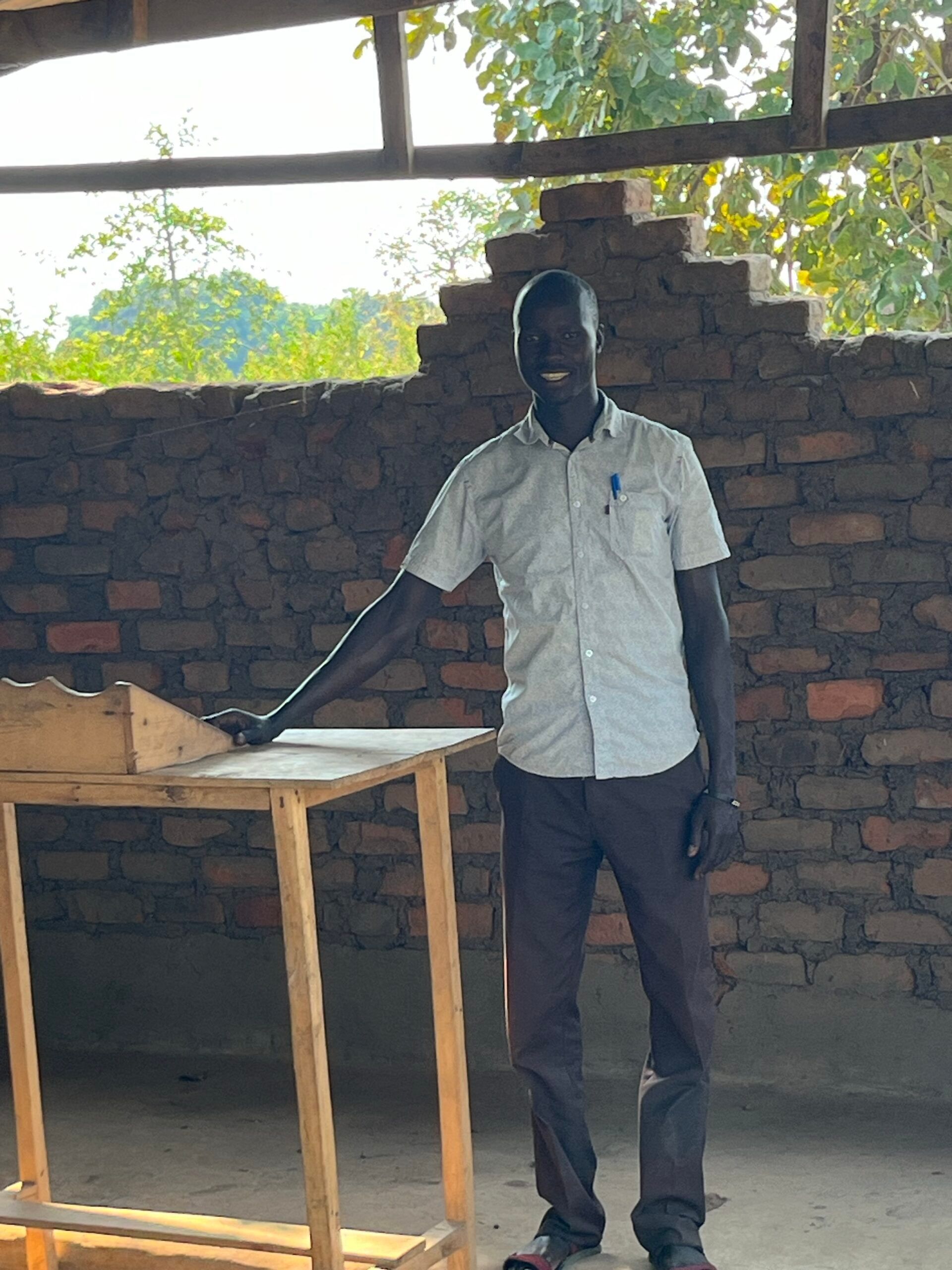 Man stands next to wooden table. He is smiling and wearing a light shirt and dark pants in a brick building.