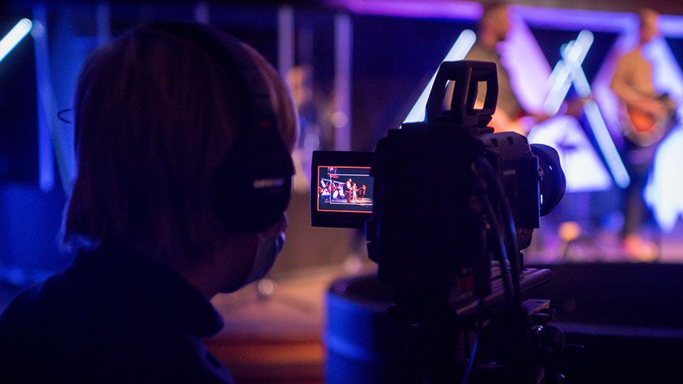 Person filming a band on stage with a video camera. The stage is lit with blue and purple lights.