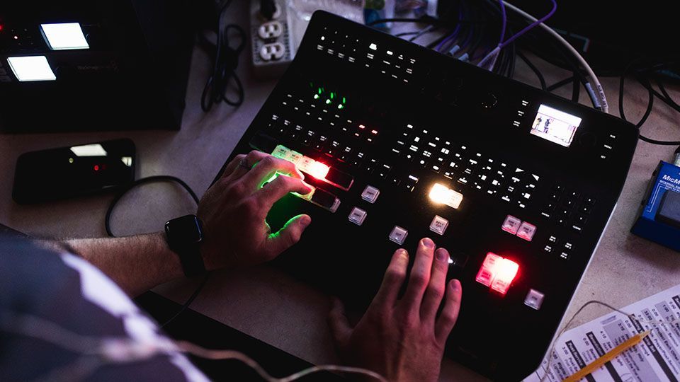 Hands operating a black video production switcher with illuminated buttons in a dimly lit setting.