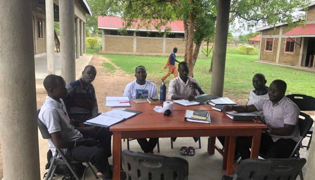 Trip 2, Post 3 Pic People seated around a table under a covered outdoor area, reviewing documents.