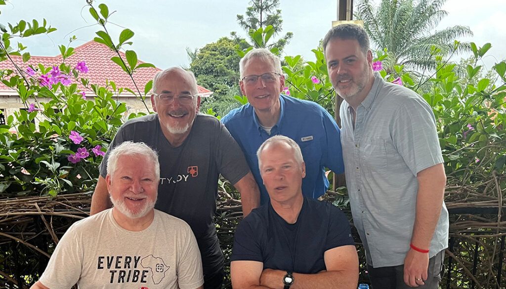 Five men pose outdoors near lush greenery, smiling.