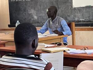 A man in a light blue shirt teaches in a classroom, facing students seated at desks.
