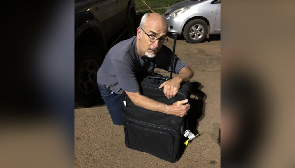 anotherday Man kneeling, hugging a large black bag next to a car.