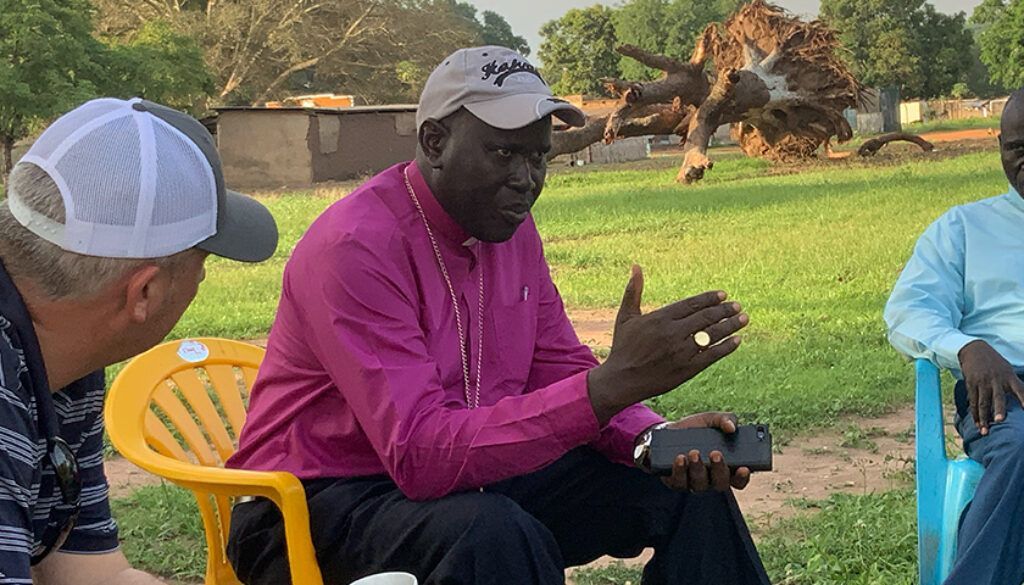 backfromafricapost Man in pink shirt gesturing, speaking to two others seated outdoors, near a fallen tree.