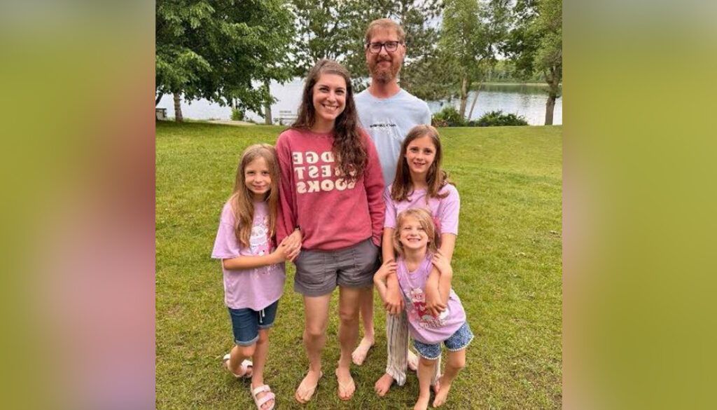 Family of five smiling, standing barefoot in a grassy outdoor setting, near trees and a body of water.