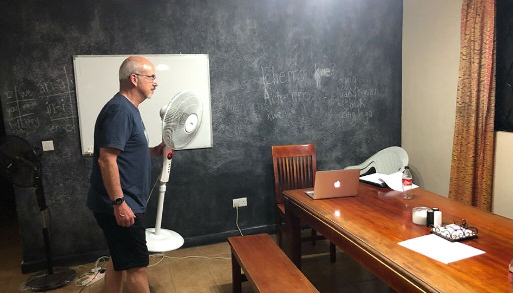 blog2-3 Man standing near a fan, pointing it toward himself. Room with a whiteboard and long wooden table.