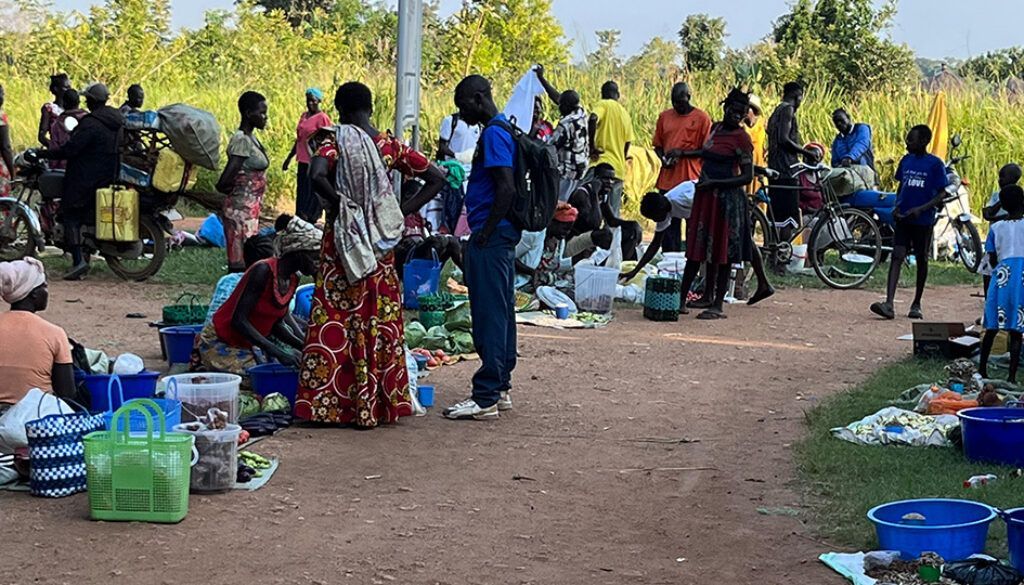 Market scene with people gathered around vendors selling goods; mostly outdoors.