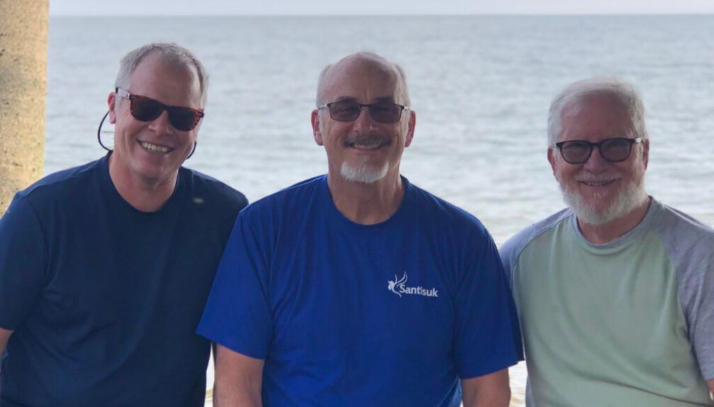 blog6 Three men wearing sunglasses smiling in front of the ocean.