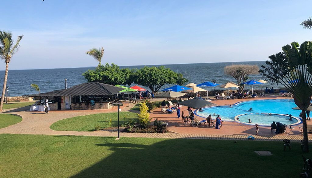 blog7 Lakeside resort with a swimming pool, people, and a thatched-roof bar. Blue water, green grass, and sunny day.