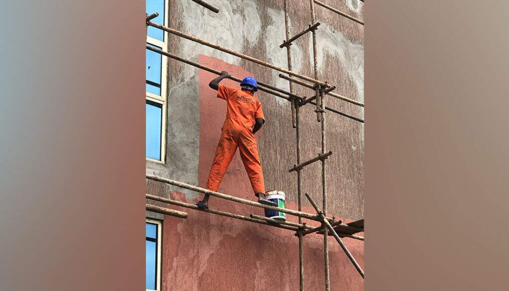 blog8 Construction worker in orange suit paints building exterior while standing on scaffolding.