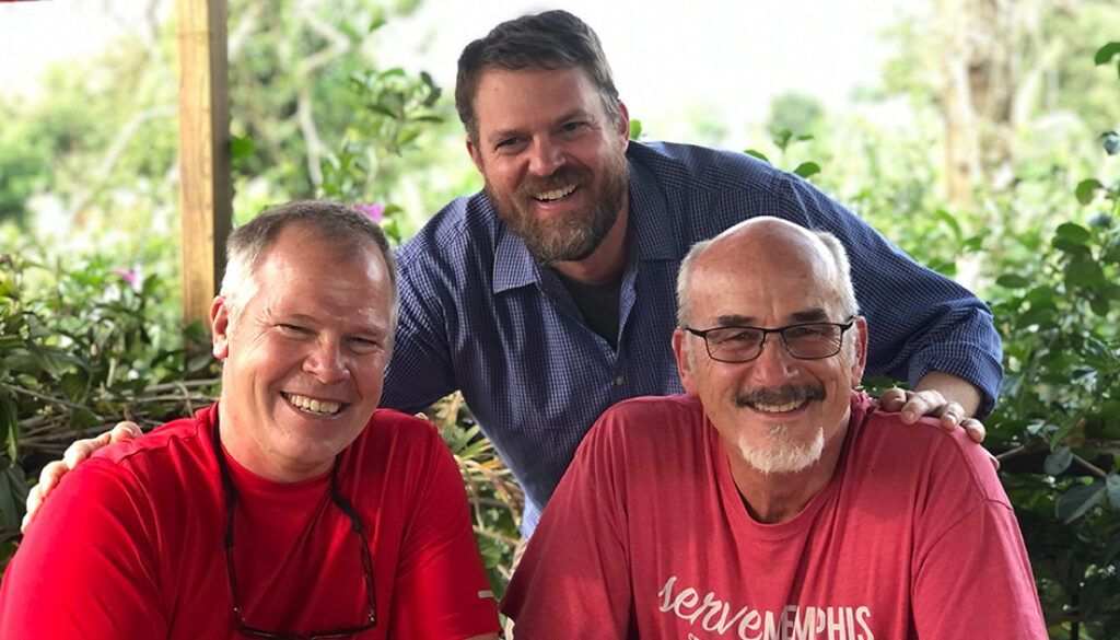 Three smiling men pose together outdoors. Two seated in red shirts, one standing behind.