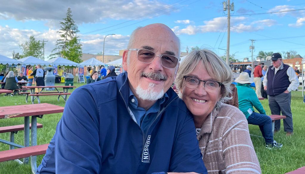 Couple smiles at camera outdoors, sitting at a picnic table with a crowd in the background.