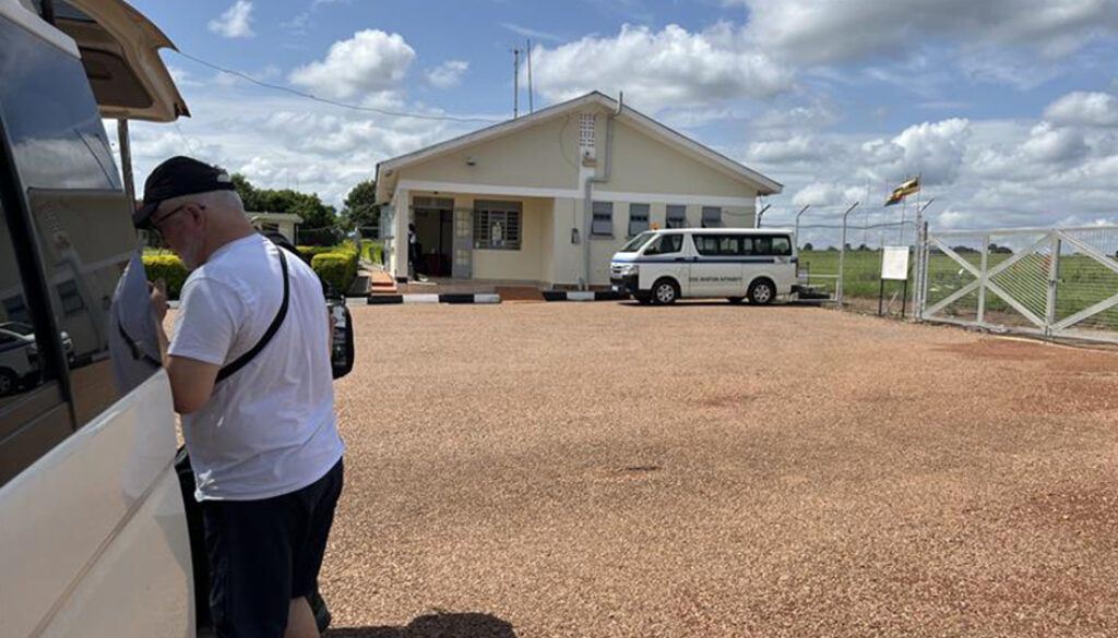 Man exiting a white van at a building with a white van parked in front. Gravel ground, fence and blue sky.