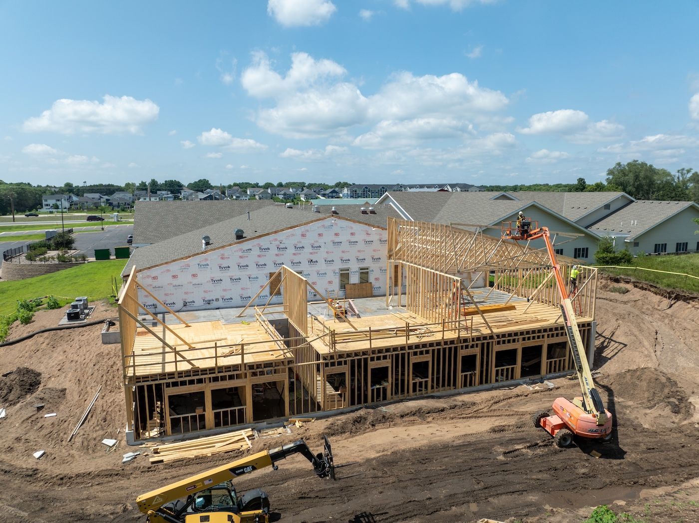 Construction site: partially built house with wooden frame, earthmoving equipment, and a lift operating on a sunny day.