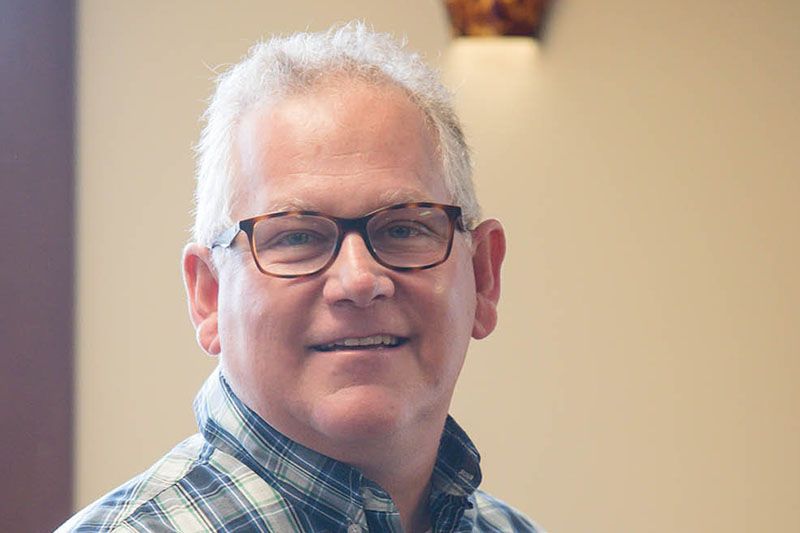 Man wearing glasses smiles, looking towards camera. He is in a room, light-skinned.