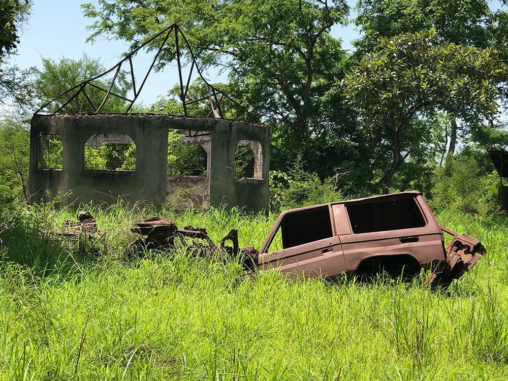 Ruined concrete building and rusted vehicle sit in overgrown grass, trees in the background.