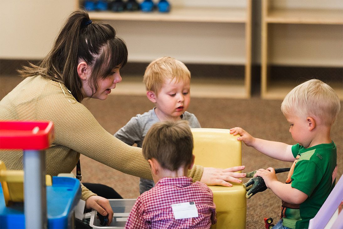 A woman helps three toddlers play with blocks in a classroom.
