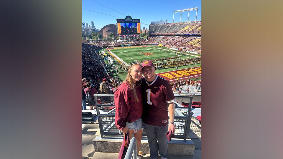 Two smiling fans in maroon shirts posing at a football stadium with the field and scoreboard behind them
