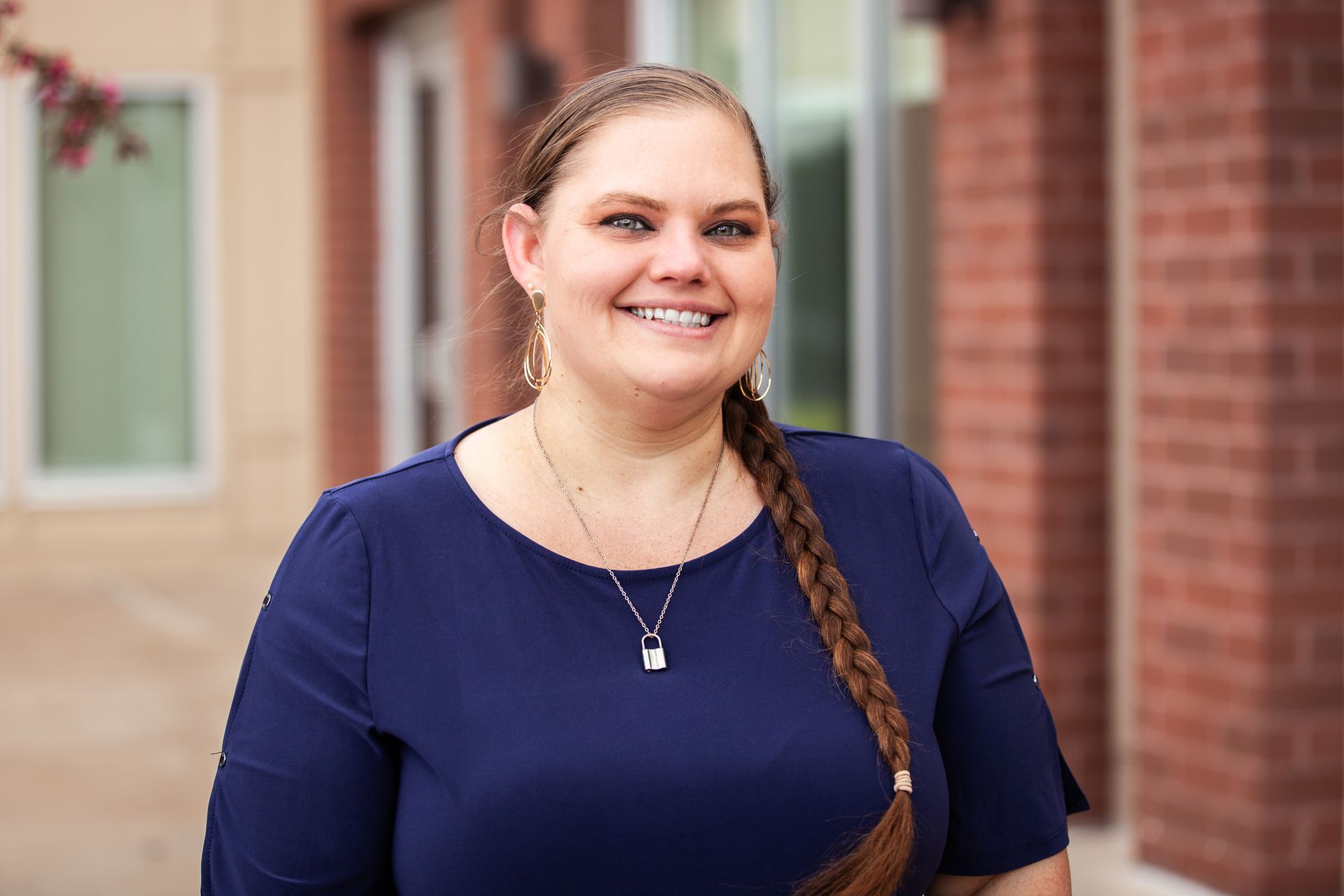 Woman with a braided ponytail smiles, wearing a navy blue top. Stands outside a brick building.
