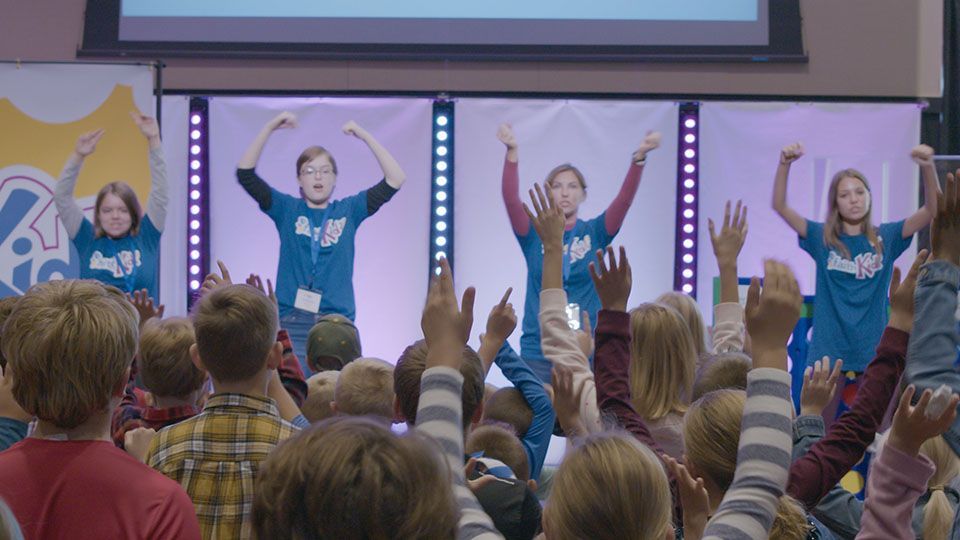 Children and four adults on stage with raised hands during a gathering in a brightly lit hall.
