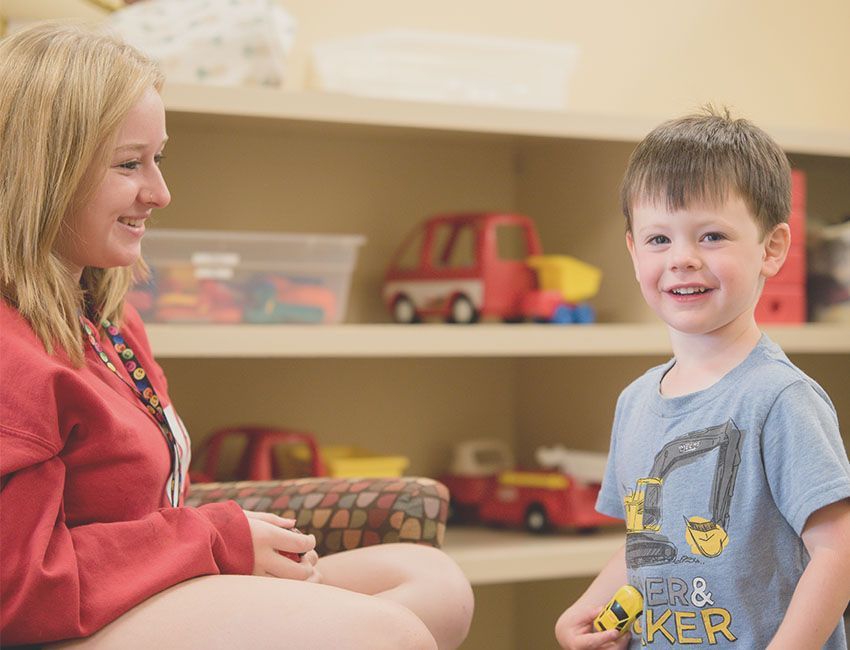 Woman in red smiles at a boy in blue, who holds a toy and smiles. Toys on shelves in background.