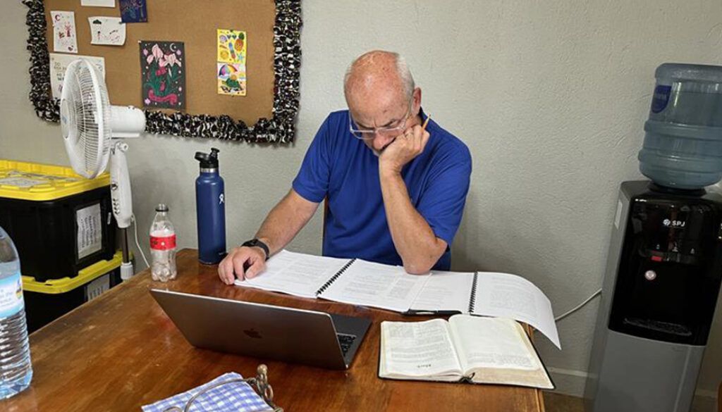 Man at desk with papers, laptop, and open book, resting head on hand.