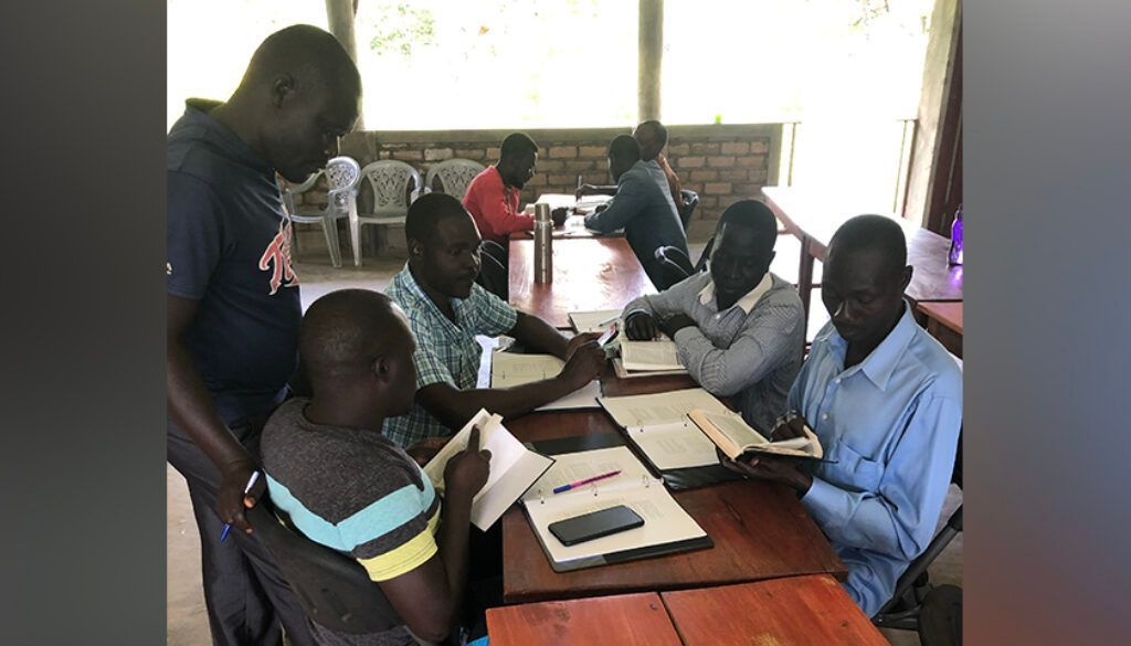 People seated around a table, reading books and writing. One man stands, looking over the shoulder of another.